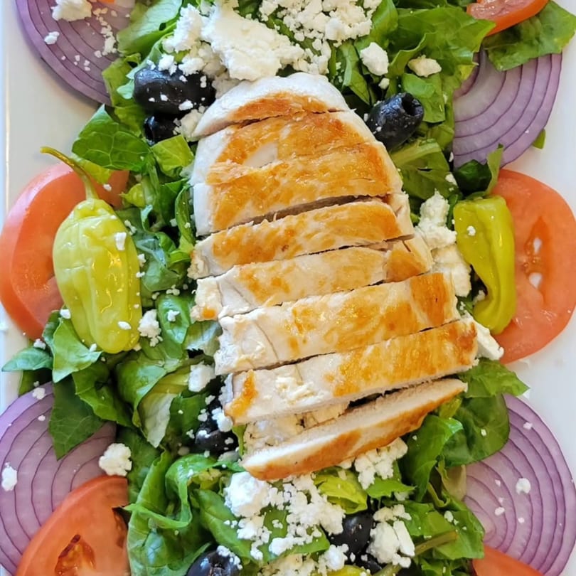 A vibrant salad with sliced grilled chicken, lettuce, tomatoes, red onion, olives, feta, and peppers.