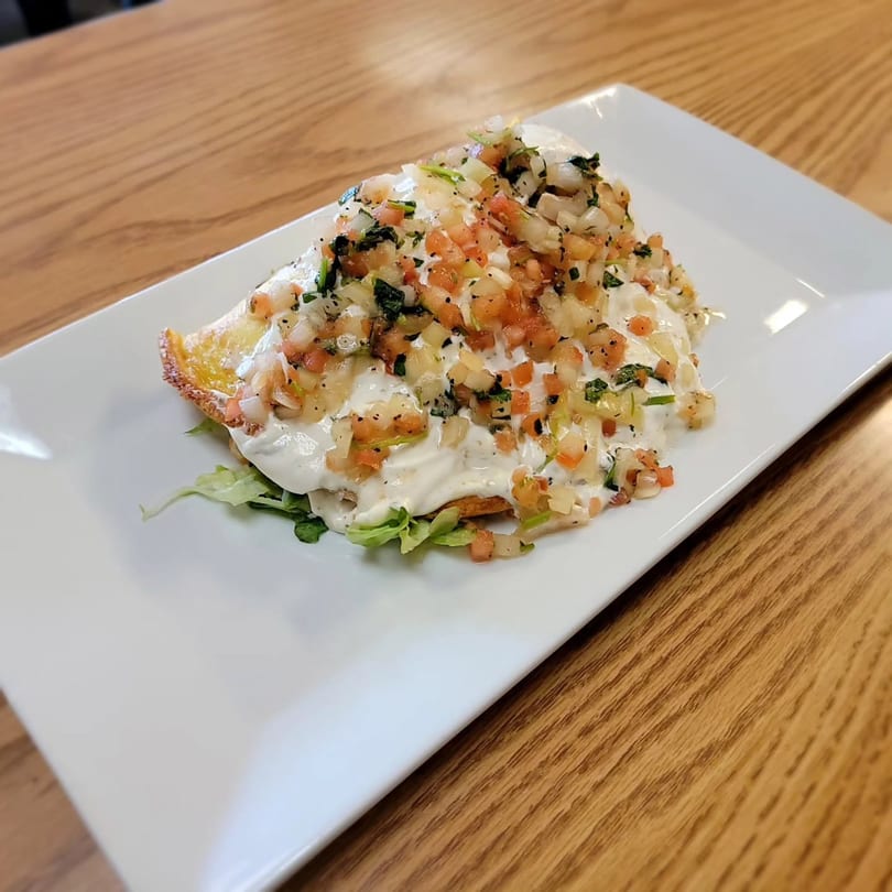 A golden-brown fried tortilla topped with white sauce, diced tomatoes, onions, and herbs.