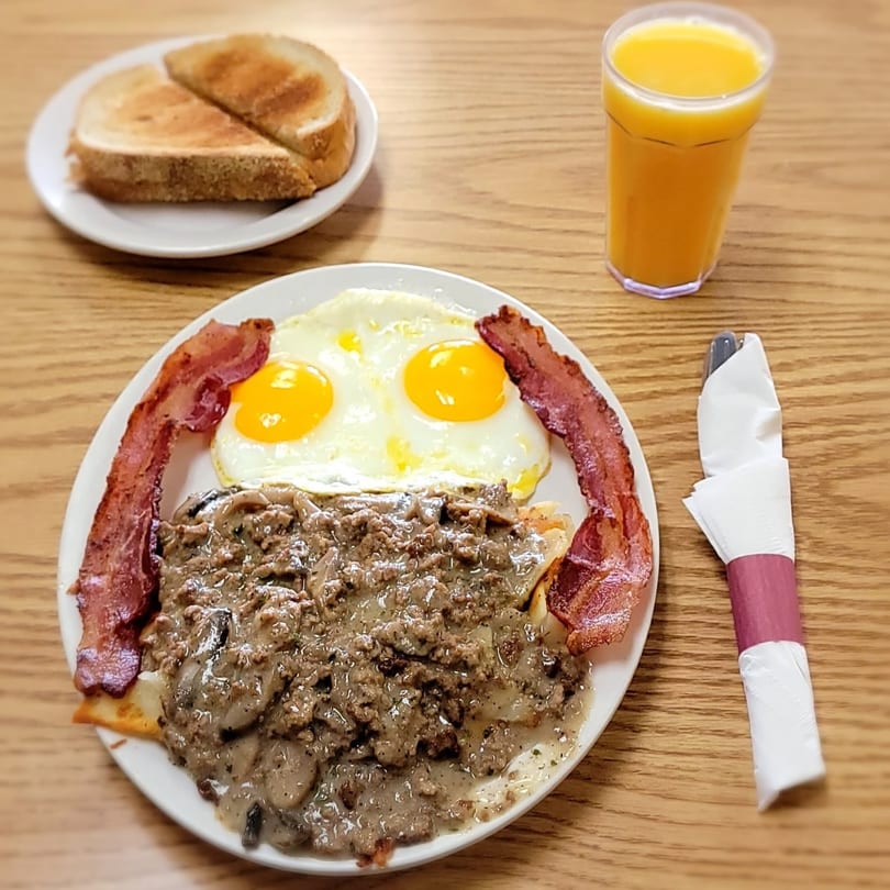 Breakfast plate with eggs, bacon, gravy, toast, and orange juice on a wooden table.