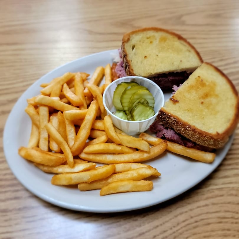 A reuben sandwich cut in half, with a side of french fries and a cup of pickle slices.