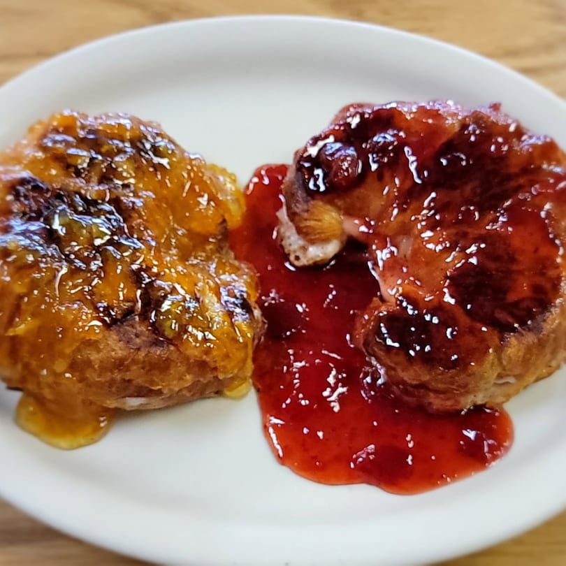 Two golden-brown pastries, one with orange glaze and the other with red jam, on a white plate.